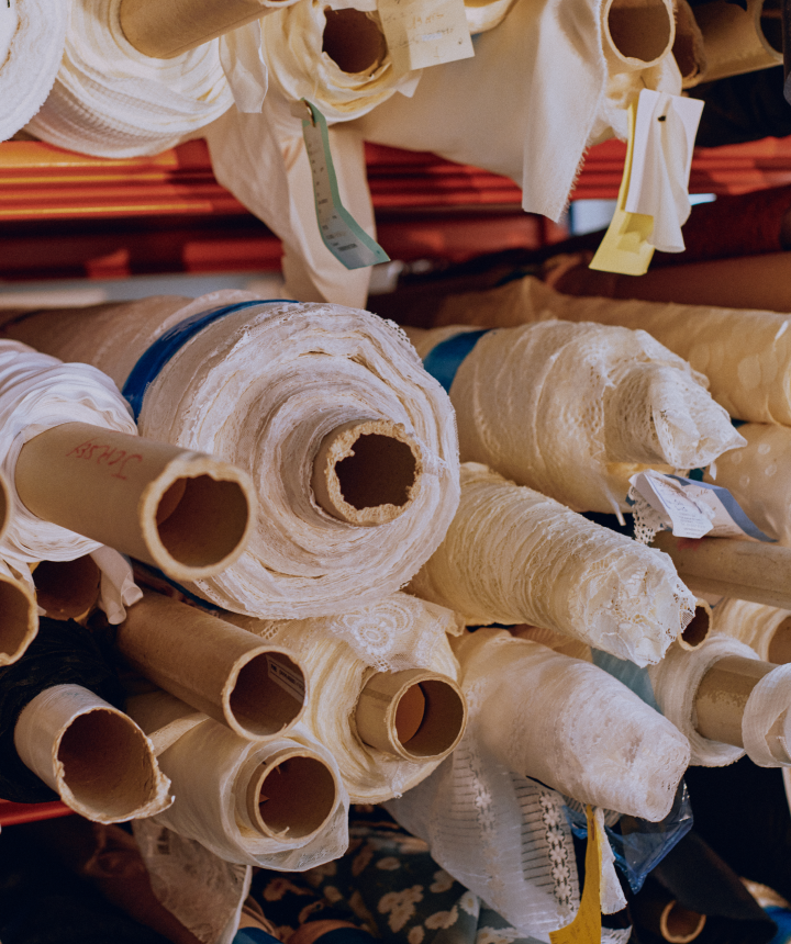 Close-up of numerous rolls of white and cream fabric, some with tags, in a textile workshop or warehouse, suggesting raw materials for fashion design or manufacturing.