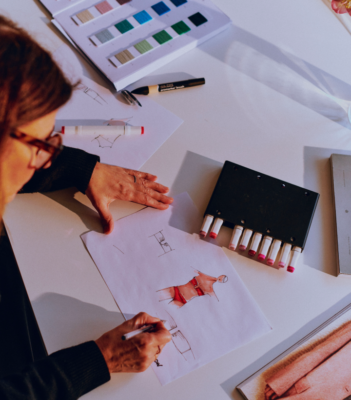 Fashion designer drawing a sketch of a swimsuit at a desk with markers and magazines.