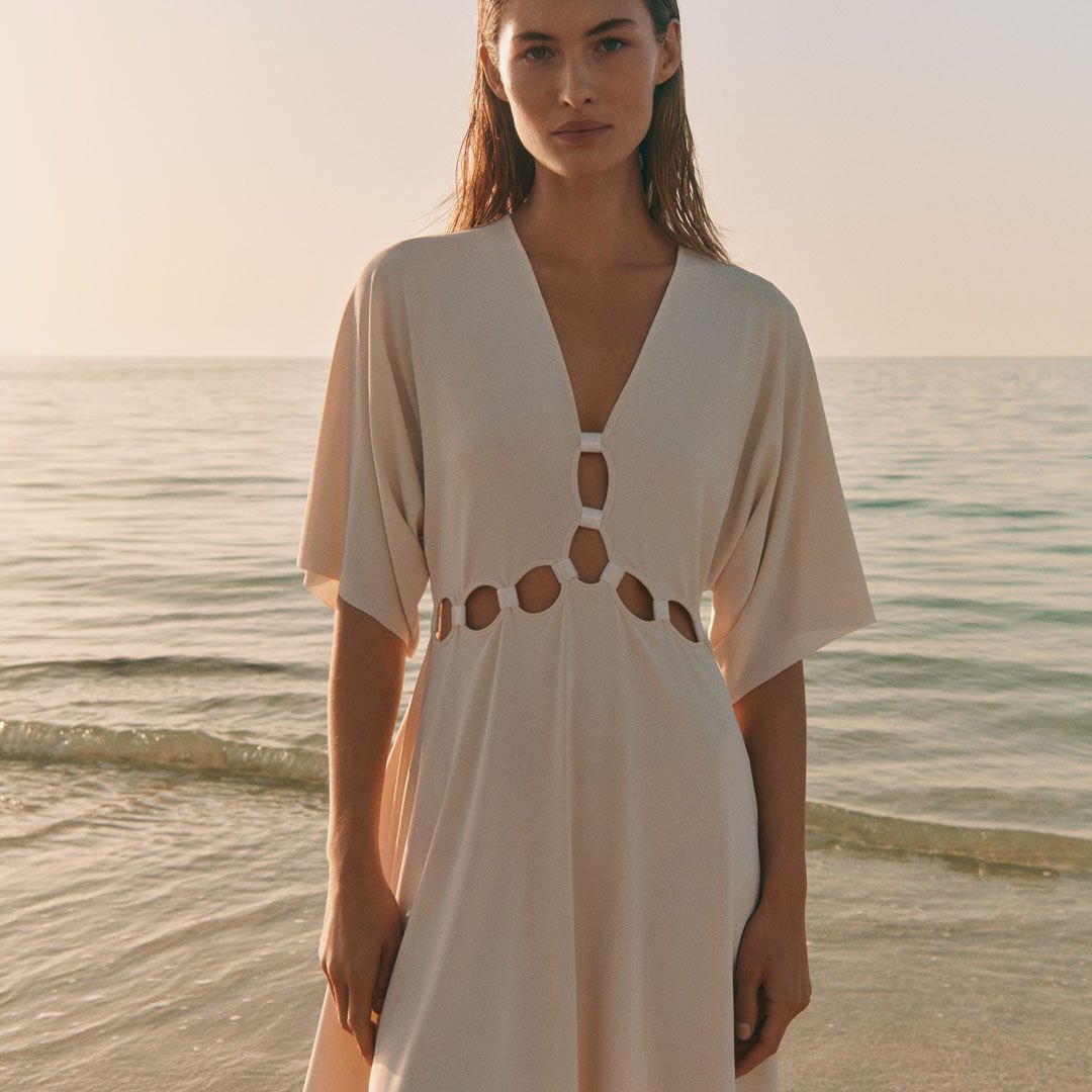 Woman in a beige dress with cut-out details standing by the sea at sunset.