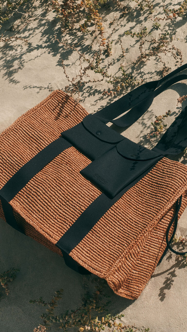 Cabas tress&eacute; marron avec anses noires et pochettes noires attach&eacute;es, pos&eacute; sur le sable avec lumi&egrave;re naturelle et ombres.