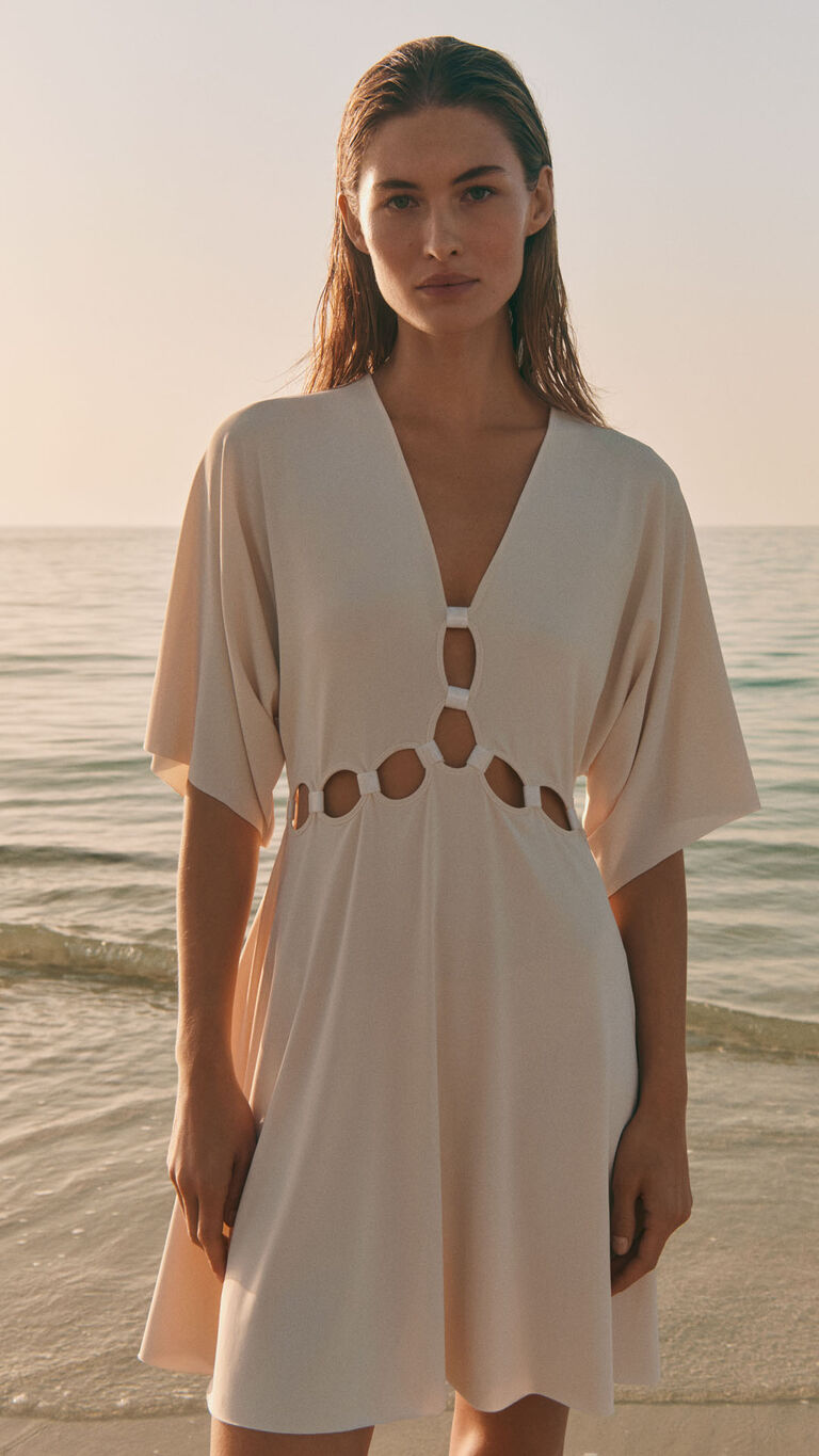 Front view of a woman wearing a light beige dress with cut-out details at the waist, standing by the sea at sunset.