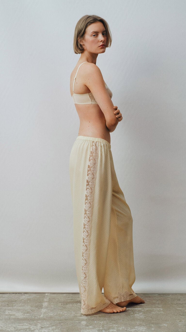 Una mujer con sujetador beige de encaje y pantal&oacute;n color crema est&aacute; de perfil.