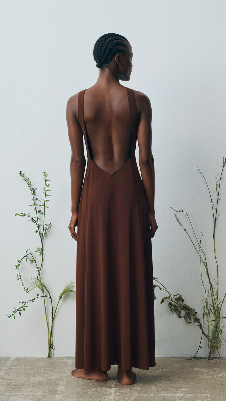 Model photographed from the back wearing a long brown dress with a deep open-back design and flowing silhouette.