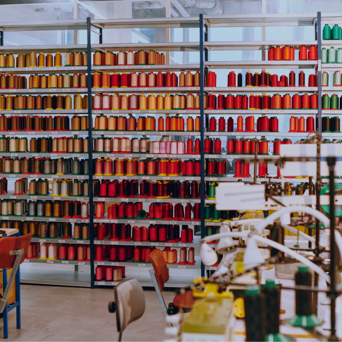 Extensive wall of shelves filled with colorful sewing thread spools in a textile factory or workshop, indicating materials for sewing, embroidery, or industrial garment production.